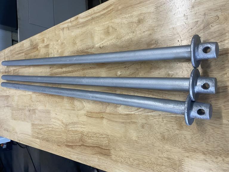 Three galvanized barrier pins laid side by side on a workbench, showing full-length shafts and flanged bases
